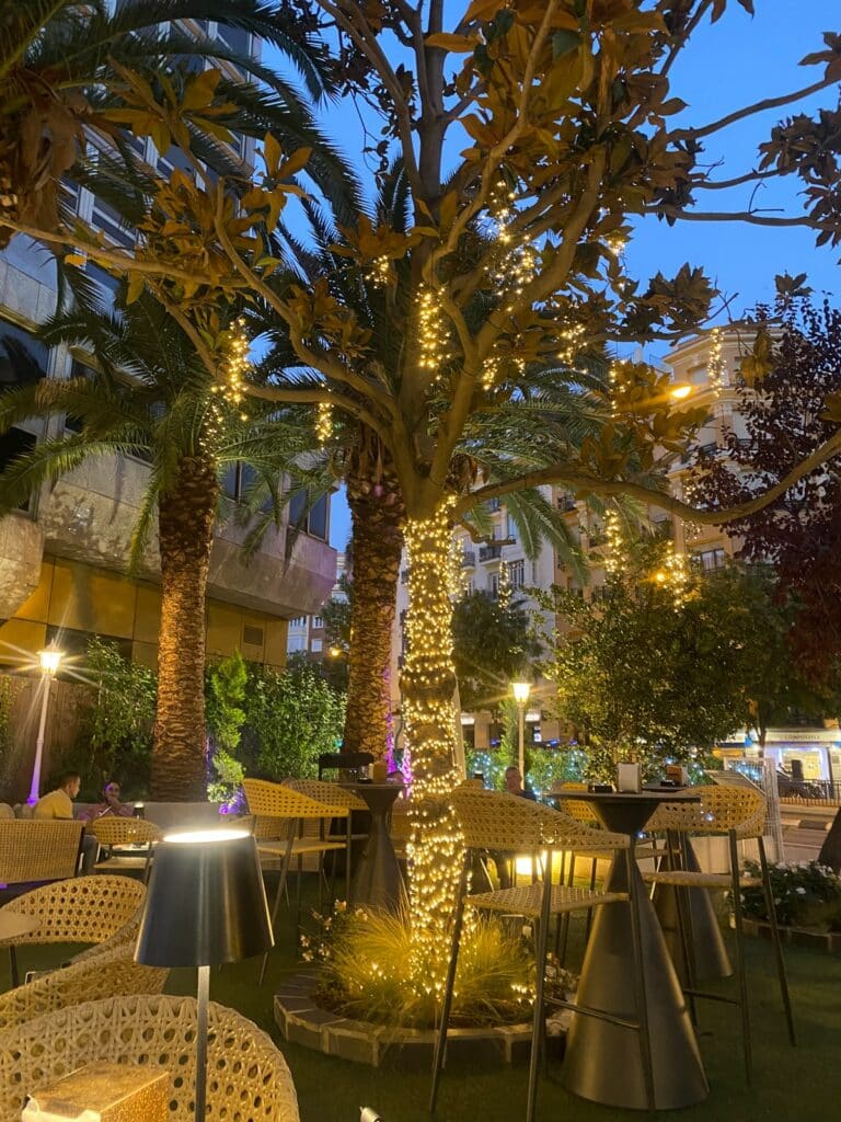 La terraza del hotel, vestida con mesas y sillas altas y detalles de iluminación como lámparas y luces en las palmeras.